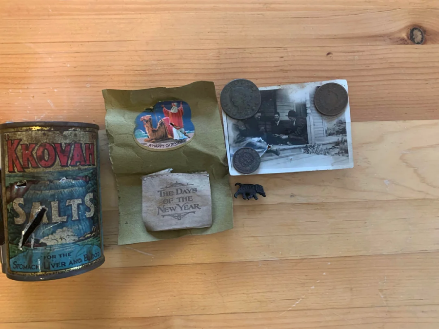 A collection of vintage items on a wooden surface, including a tin labeled "Kkovah Salts," a greeting card, a small stone with "The Days of the New Year" written on it, coins, a black animal figurine, and an old black-and-white photo.