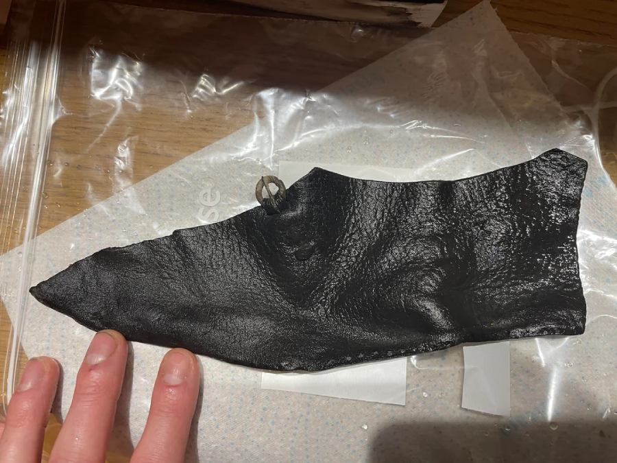 A hand holds a thin, black, shiny piece of textured material resembling leather, placed on a transparent plastic sheet with paper underneath on a wooden surface.