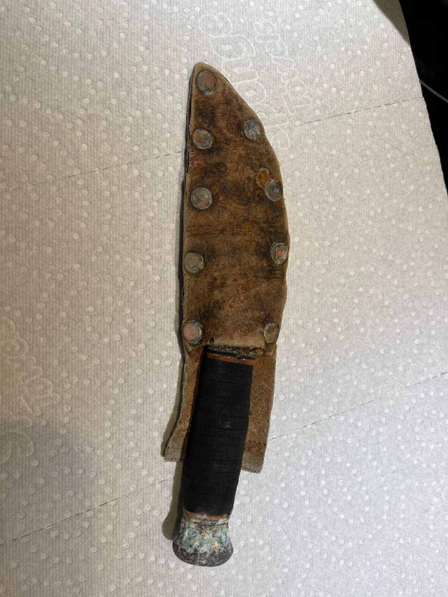 A knife with a worn leather sheath secured by metal rivets, and a handle wrapped in dark material, rests on a textured white surface.