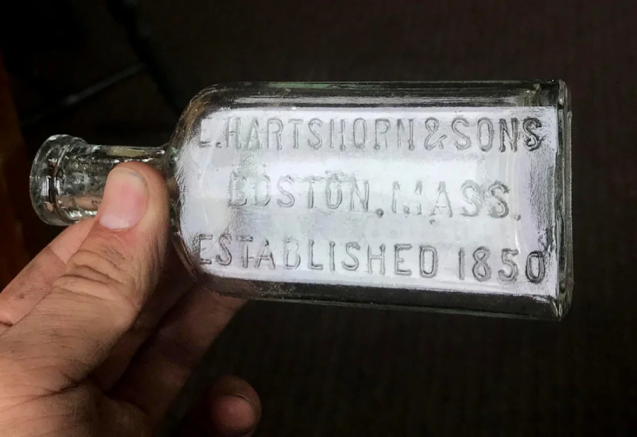 A hand holds a clear glass bottle embossed with the text: “E. Hartshorn & Sons, Boston, Mass., Established 1850.” The background is out of focus.