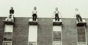 Four people sit separately on the edges of concrete ledges above windows on a brick building, appearing as if suspended high in the air. The image is in black and white.
