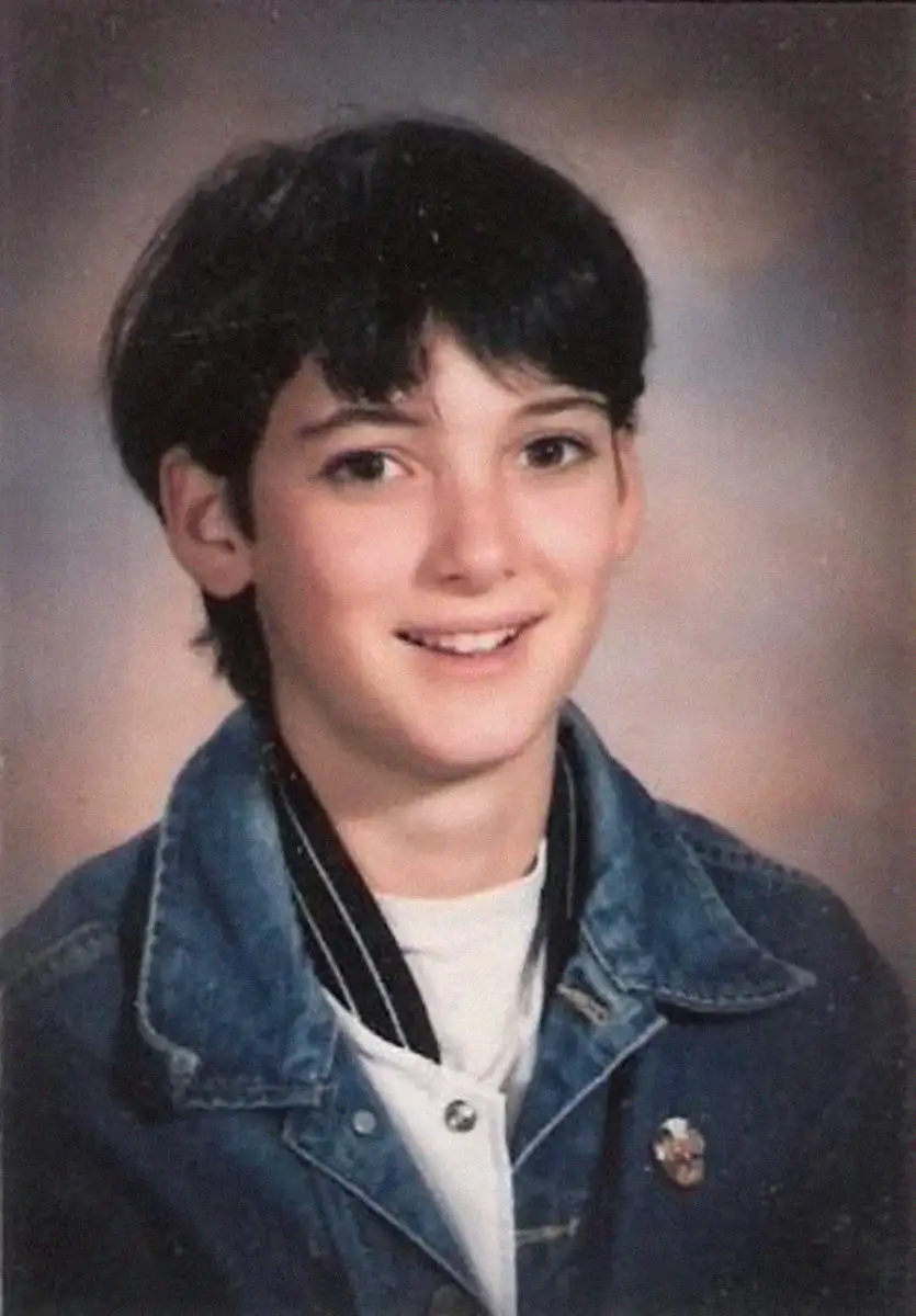 A young person with short dark hair, wearing a denim jacket over a white shirt and a striped collar, smiles at the camera in a school portrait with a softly blurred brown background.