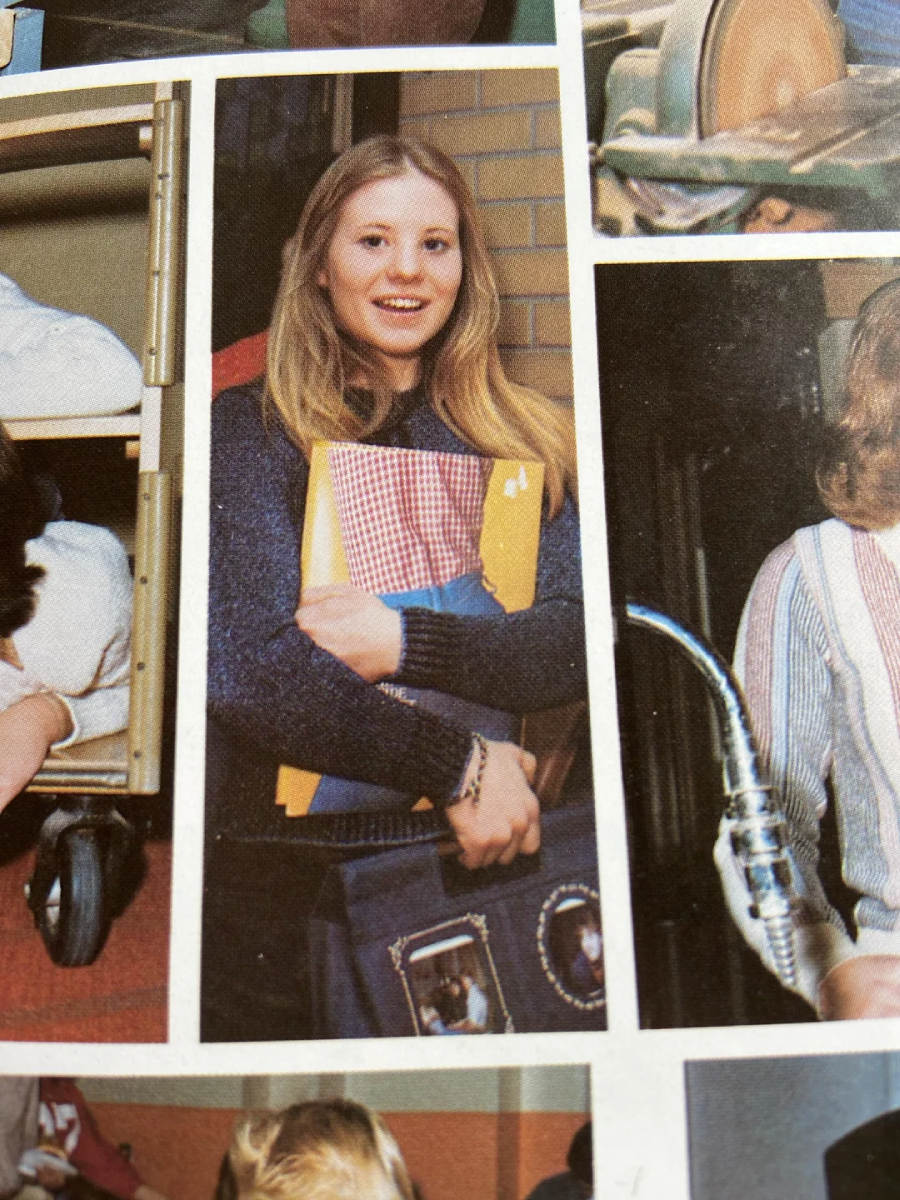 A teenage girl with long blonde hair smiles while holding books and folders. She is wearing a navy blue sweater and stands in front of a brick wall, posing for a photo.