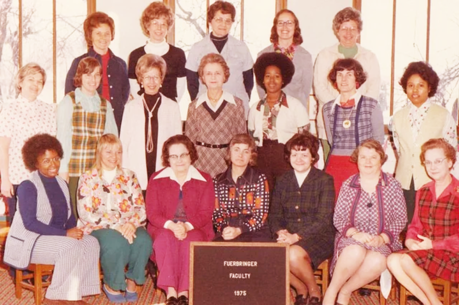A group of women in colorful 1970s clothing pose indoors for a faculty photo, with a sign in front reading "Fuehrminger Faculty 1975"; some are seated, others stand behind them in rows.