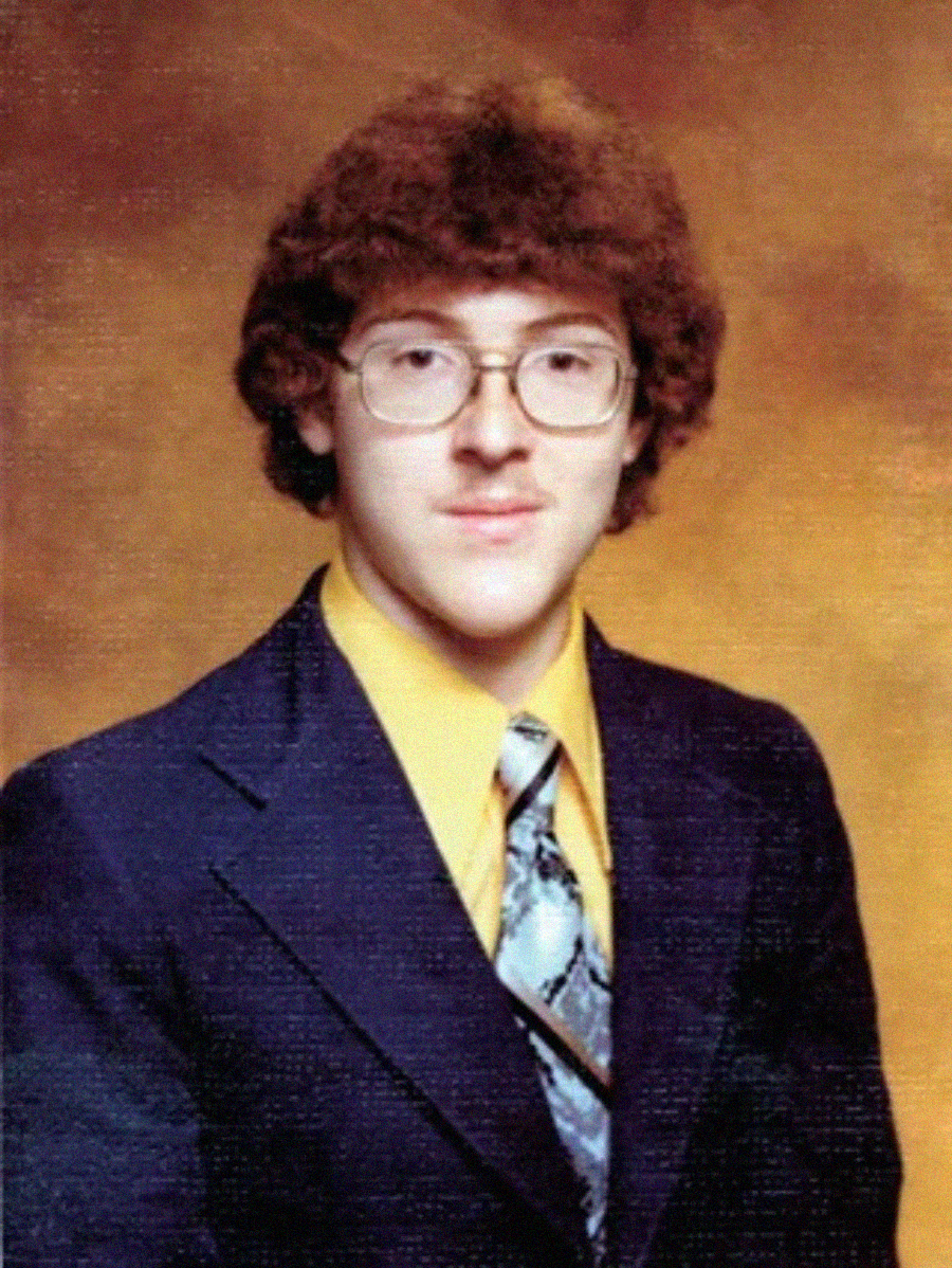 A young man with curly brown hair, large glasses, and a faint mustache wears a yellow shirt, patterned tie, and dark suit jacket, posing in front of a brown mottled studio backdrop.