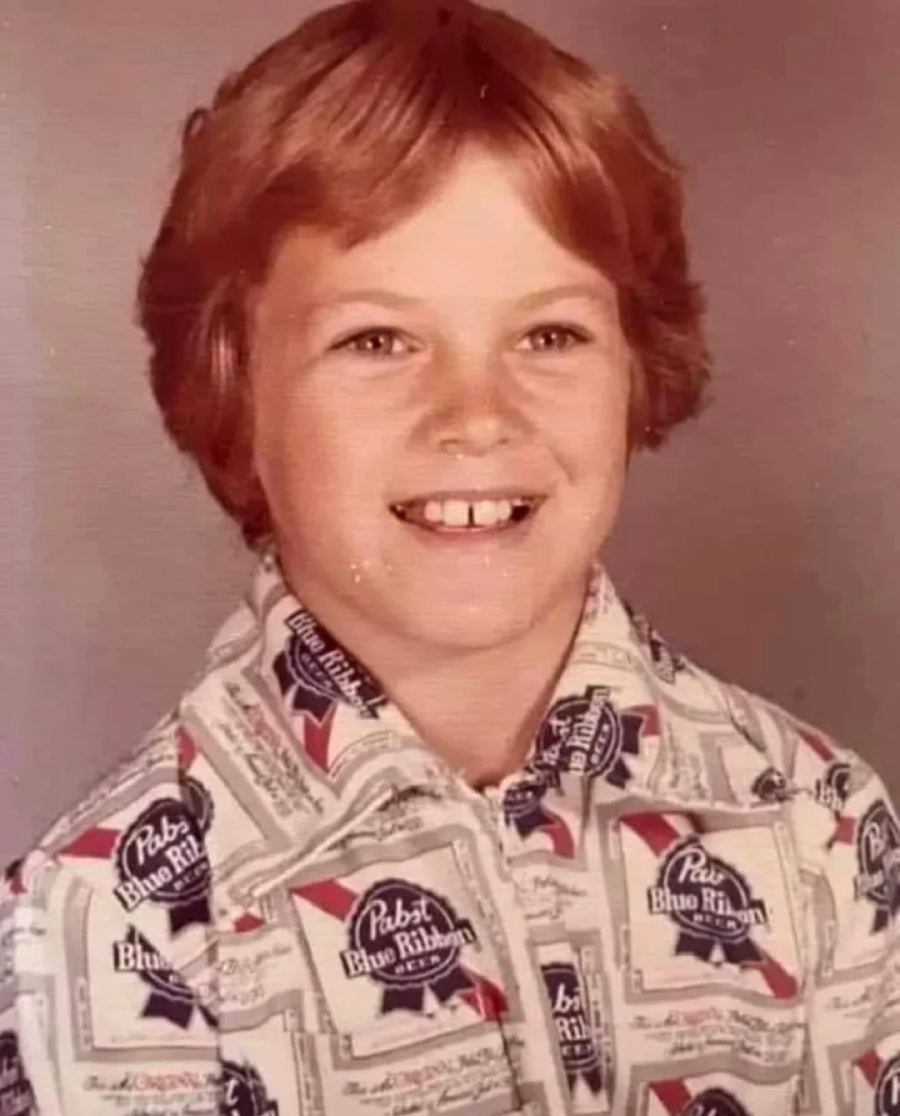 A smiling child with short, light brown hair wears a collared shirt covered in a repeating Pabst Blue Ribbon beer logo pattern, posing for a vintage-style school photo.