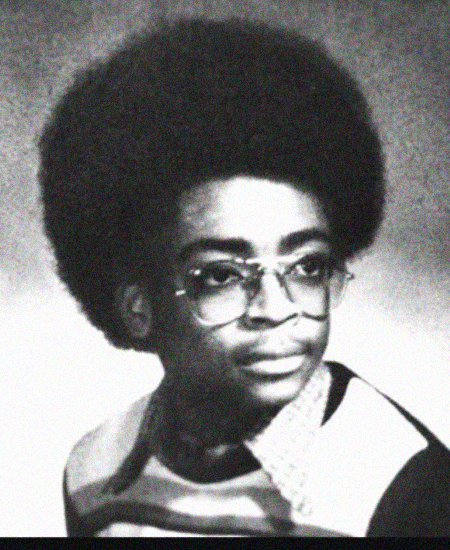 Black and white yearbook-style portrait of a young person with an afro hairstyle, wearing glasses and a collared shirt with a patterned sweater. The background is a neutral gray.