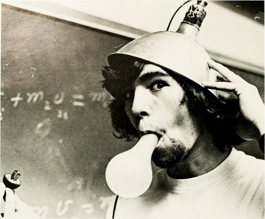 A young man with long hair wears a metal bowl with wires as a hat and blows up a balloon with his mouth, standing in front of a chalkboard covered in math equations.