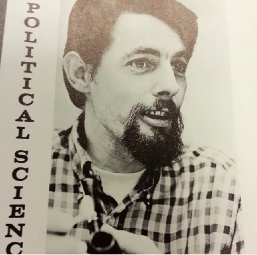 Black and white photo of a man with a mustache and goatee, wearing a plaid shirt, holding a pipe. The words "POLITICAL SCIENCE" are written vertically along the left side of the image.