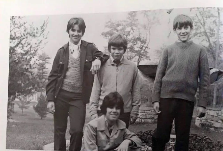 Four boys pose outdoors near a large fountain or sculpture, smiling at the camera. Three stand behind, while one crouches in front. The photo is black and white, and trees are visible in the background.