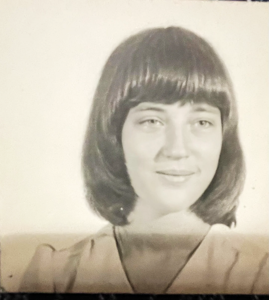 A black-and-white portrait of a young woman with short, straight dark hair and bangs, wearing a light-colored blouse, smiling softly while looking slightly to the side.
