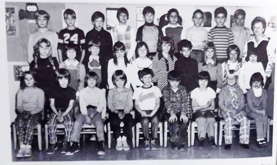 A black-and-white photo of a classroom group with about 30 children and one adult, arranged in three rows, some standing and others seated, posing and smiling for the camera in a school setting.