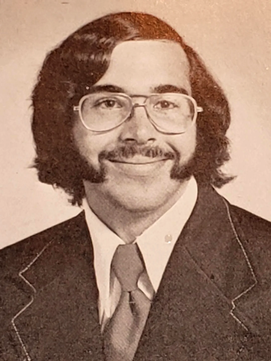 A young man with thick glasses, a mustache, and prominent sideburns smiles gently. He is wearing a suit jacket, dress shirt, and tie. The photo is in black and white and appears to be a vintage portrait.