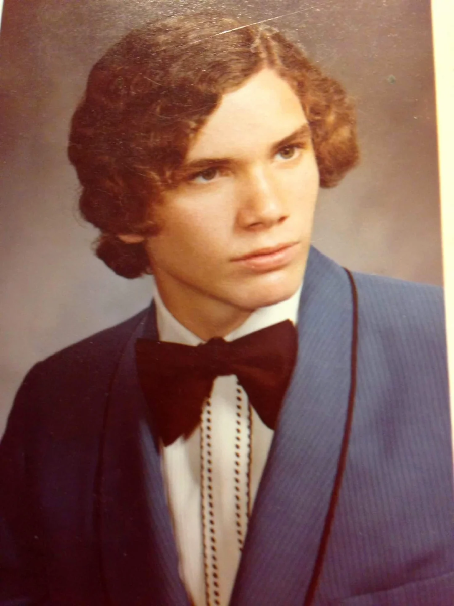 A young man with curly brown hair wearing a blue suit, white shirt, and large black bow tie poses for a formal studio portrait against a neutral background.