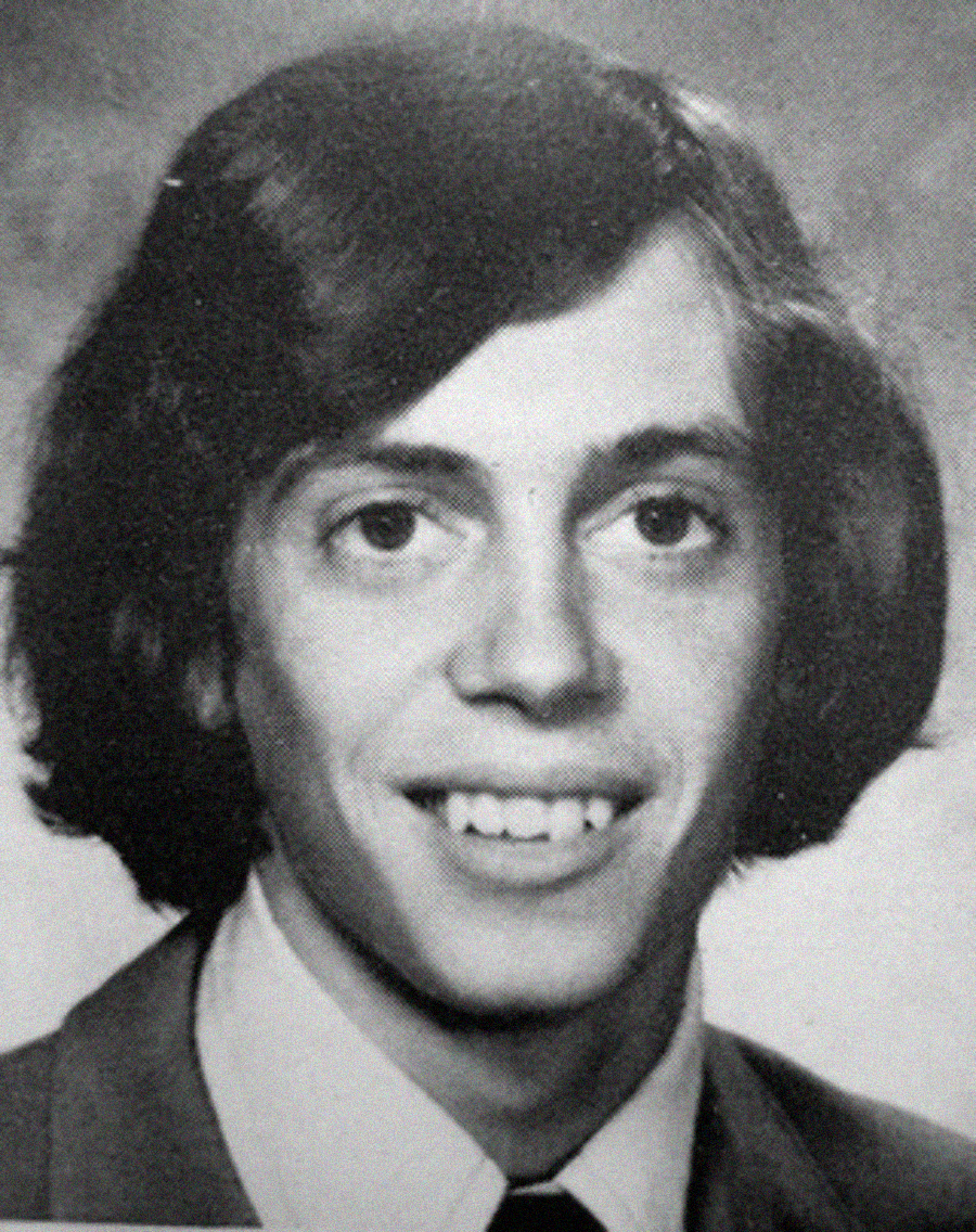 Black-and-white yearbook-style portrait of a young man with medium-length, dark hair, wearing a suit jacket, dress shirt, and tie, smiling at the camera against a plain background.