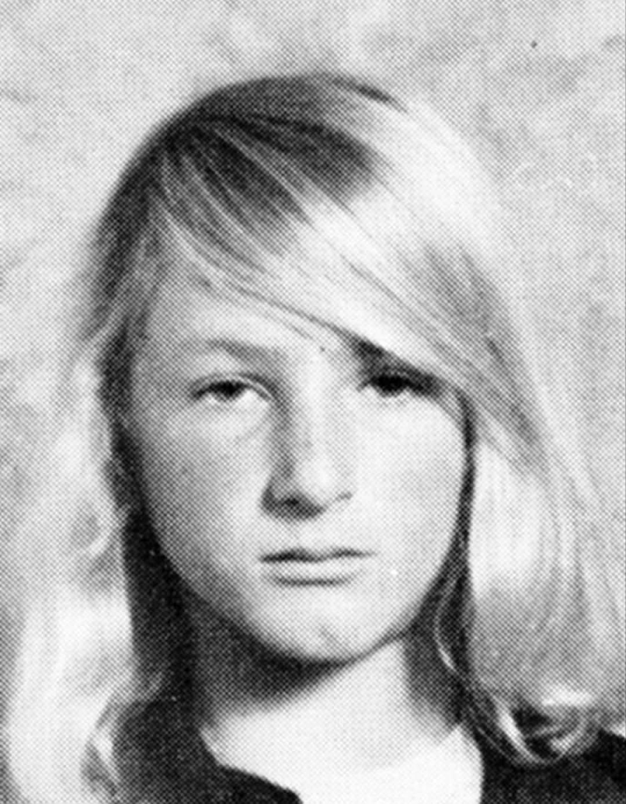 Black and white portrait of a young person with long, straight, light-colored hair, looking directly at the camera with a neutral expression. The background is plain and slightly textured.