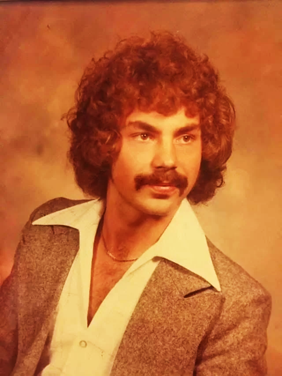A man with curly hair and a mustache wears a gray suit jacket over a wide-collared white shirt. The background is a warm, faded brown. The photo has a vintage look.