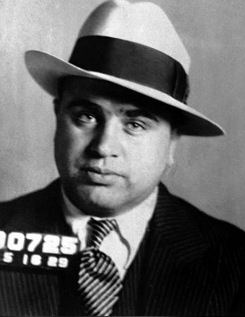 Black-and-white mugshot of a man wearing a white fedora, dark striped suit, and tie. A portion of a number plate is visible at the bottom left. The man looks directly at the camera with a neutral expression.