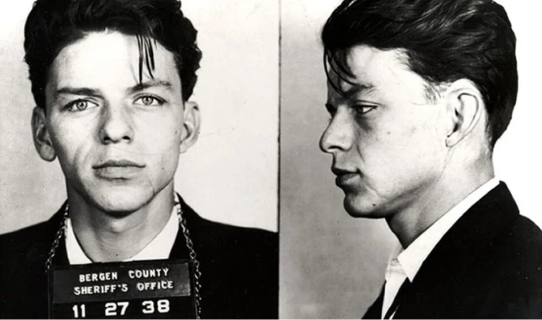 Black and white mugshot photo showing a young man in a suit, facing forward and in profile, holding a sign reading "Bergen County Sheriff's Office 11-27-38.