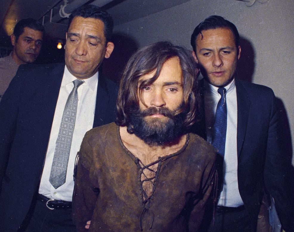 A man with long hair and a beard, wearing a brown shirt, is escorted by two men in suits and ties in what appears to be a dimly lit hallway.
