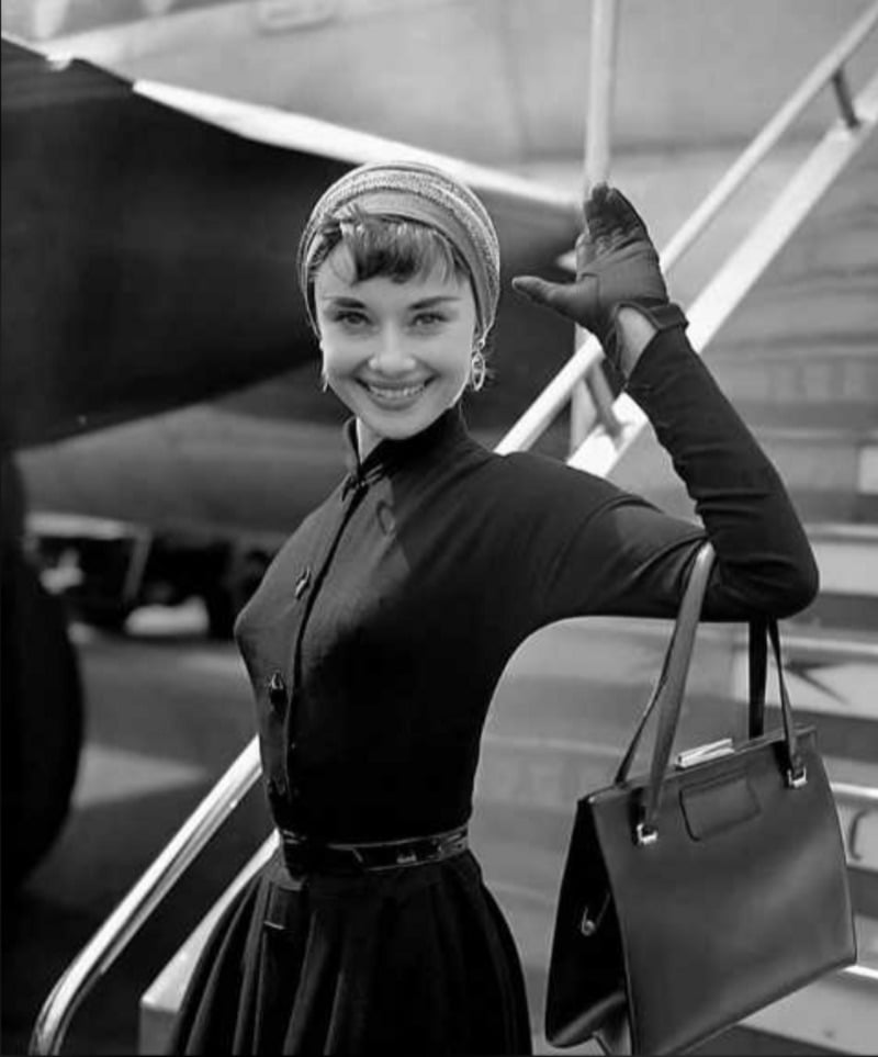 A stylish woman in a dark dress, headscarf, and gloves smiles and waves while standing beside airplane steps, holding a handbag. The image is in black and white.