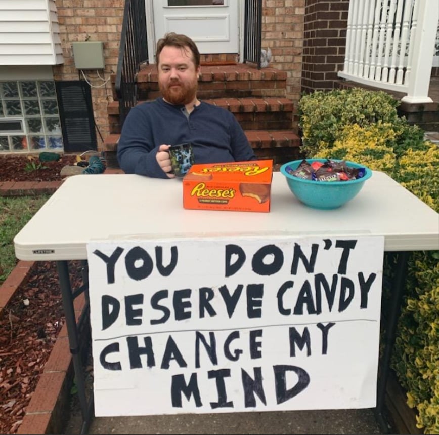 A man with a mug sits at a table outside with a large sign that reads, "You don’t deserve candy. Change my mind." The table has a Reese's box and a bowl of assorted candy.
