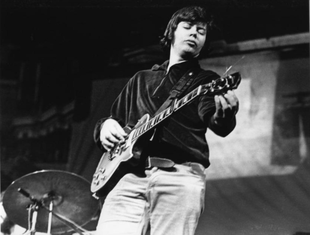 A black-and-white photo of a man playing an electric guitar on stage, with his eyes closed and head tilted. A drum set is visible in the background. The image captures a moment of concentration and musical performance.