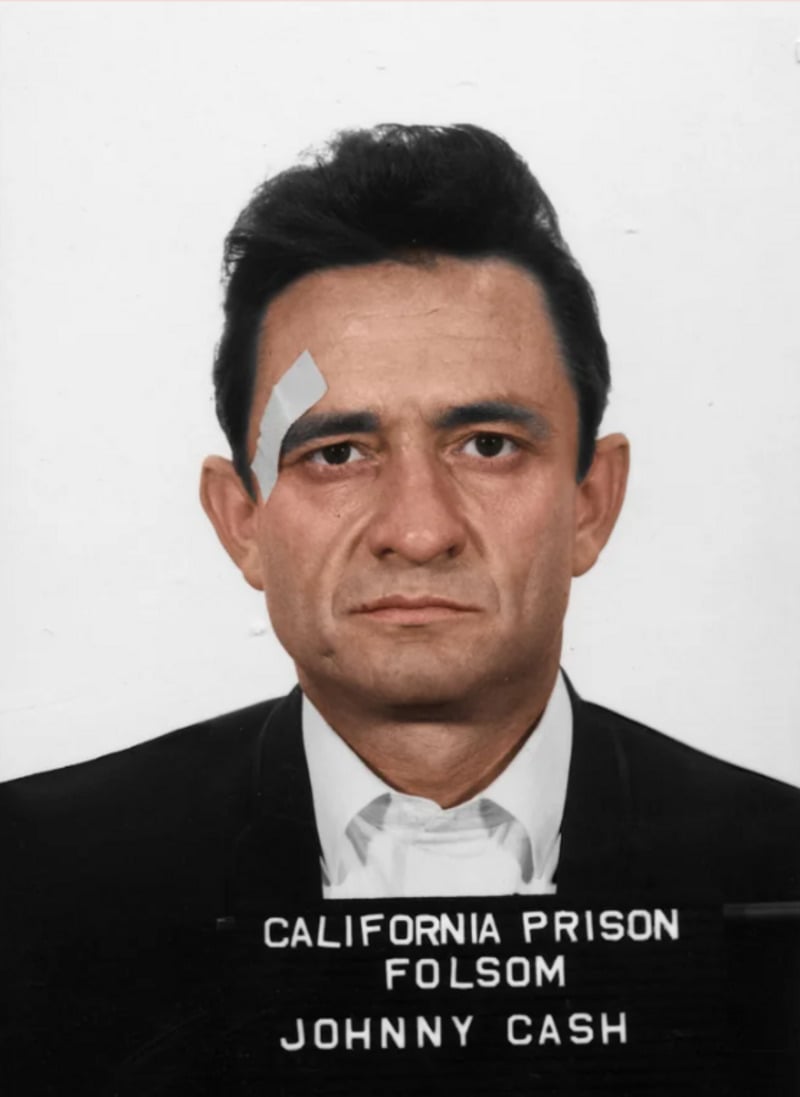 A man with dark hair and a serious expression, wearing a suit and a bandage above his left eyebrow, poses for a prison mugshot labeled "California Prison Folsom Johnny Cash" on a white background.