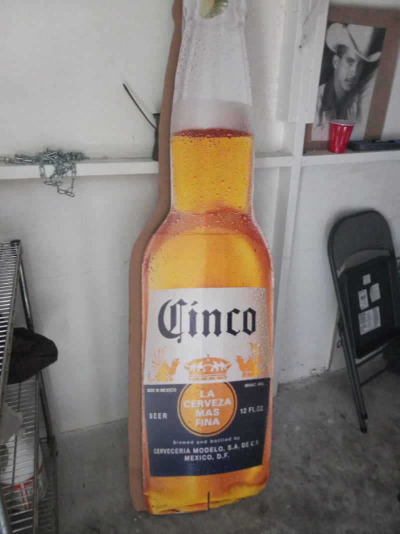A large cardboard cutout of a Cinco beer bottle stands against a wall in a garage, with a metal shelf on the left, a folding chair, a red cup, and a black-and-white photo on the right.