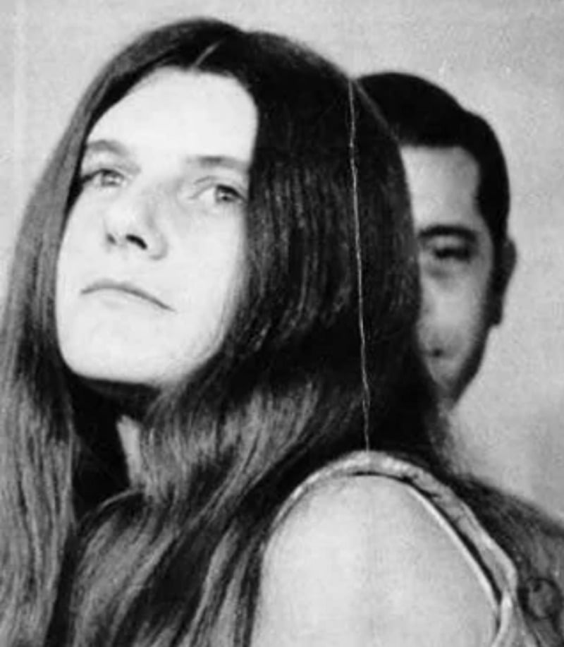 Black and white photo of a woman with long straight hair looking upward, with a man partially visible in the background, slightly out of focus.