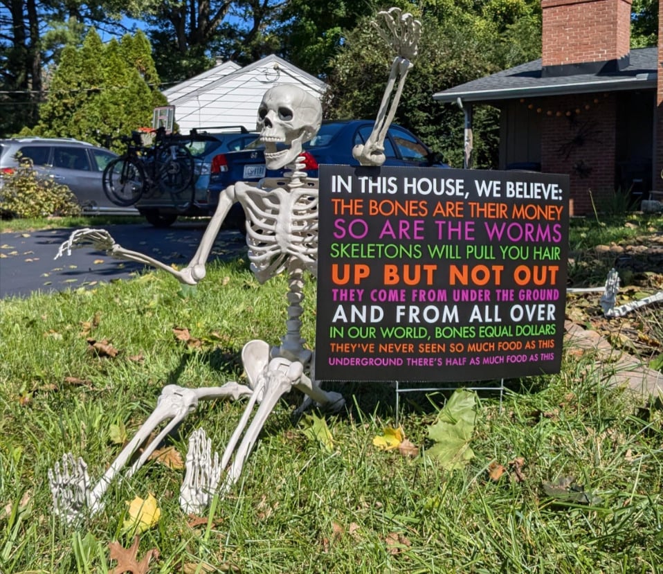 A plastic skeleton sits on a lawn by a driveway, holding a colorful sign that humorously references a song from the TV show "I Think You Should Leave" about skeletons and bones being money.