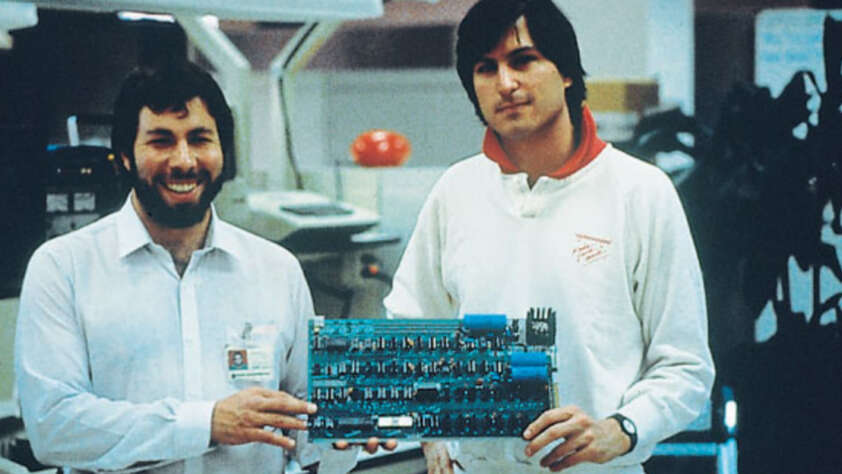 Two men stand indoors, smiling and holding an early computer circuit board. Both appear relaxed; one wears a white collared shirt and the other a white sweatshirt with a red collar. Office equipment is visible in the background.