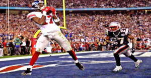 A football player in a white and red New York Giants uniform catches a pass in the end zone as a New England Patriots defender approaches during a game, with a cheering crowd in the background.
