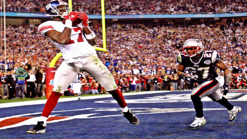 A football player in a white and red New York Giants uniform catches a pass in the end zone as a New England Patriots defender approaches during a game, with a cheering crowd in the background.