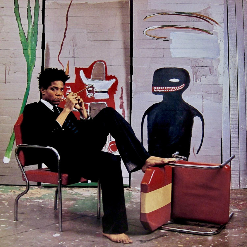 A man in a suit sits barefoot on a red chair, legs resting on another chair tipped over. Behind him are abstract paintings, including a black figure with a wide mouth and red art elements on a wooden panel wall.
