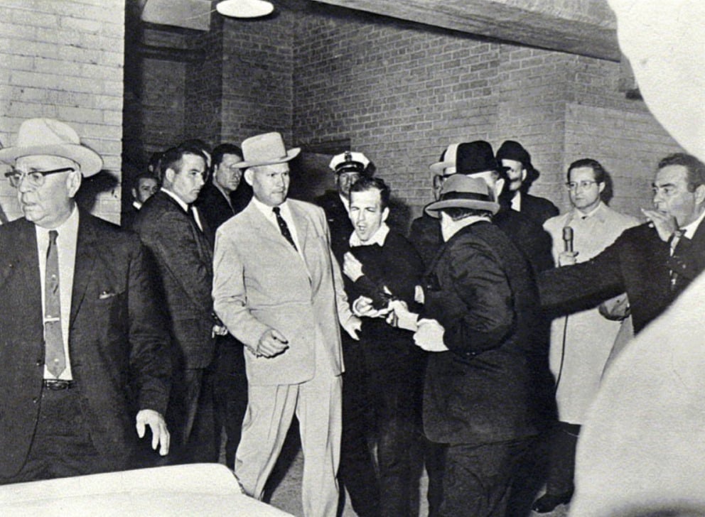 A black-and-white photo shows a chaotic scene with a man in a suit shooting another man, who appears to be in police custody, while several officers and bystanders react in shock inside a brick-walled building.