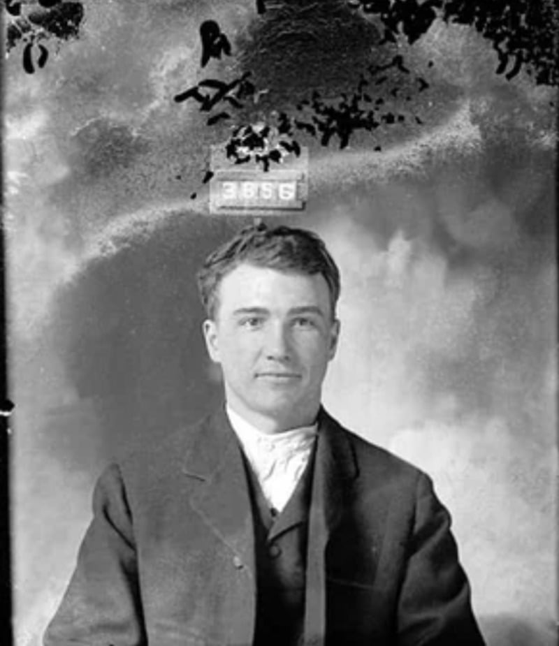 A black-and-white vintage mugshot of a man in a suit, seated with a faint smile. A placard with the number 3856 appears above his head against a blurred, textured background.