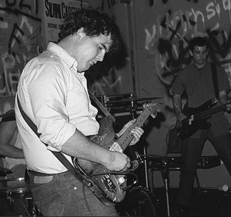 A black-and-white photo of two musicians performing onstage; one plays an electric guitar in the foreground, the other plays bass. The room has graffiti-covered walls and posters in the background.