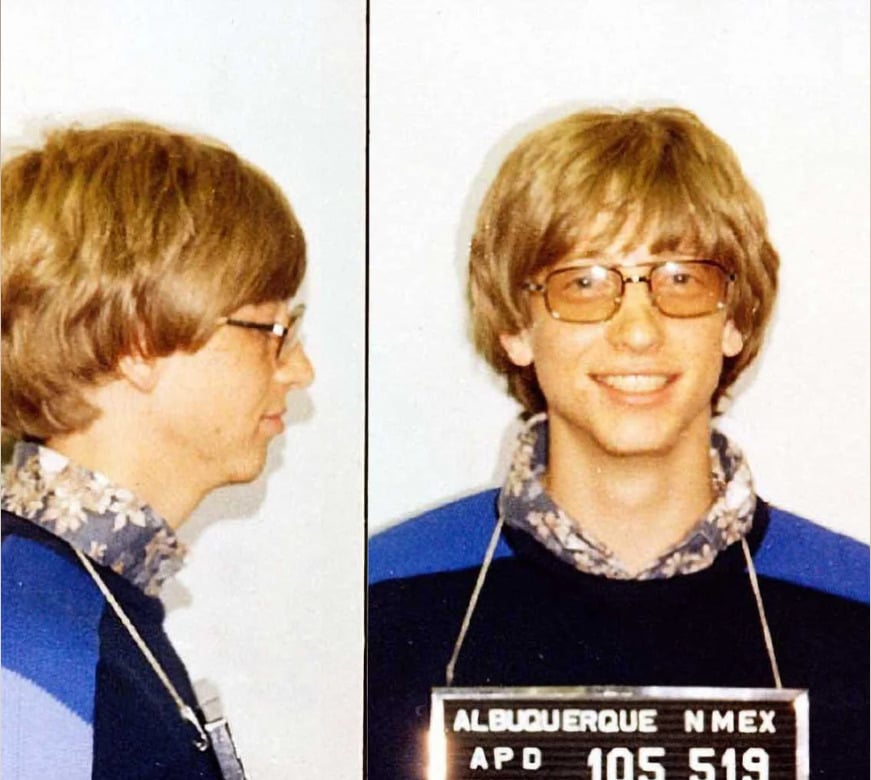 A young man with blond hair and glasses is shown in a side and front mugshot view, smiling. He wears a patterned shirt and sweater. A placard reads "ALBUQUERQUE NMEX APD 105 519.