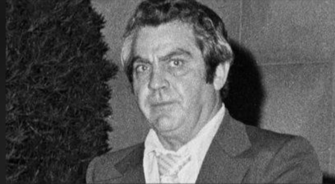 Black-and-white photo of a man with gray hair wearing a suit, white shirt, and tie, standing in front of a bush and looking toward the camera with a serious expression.