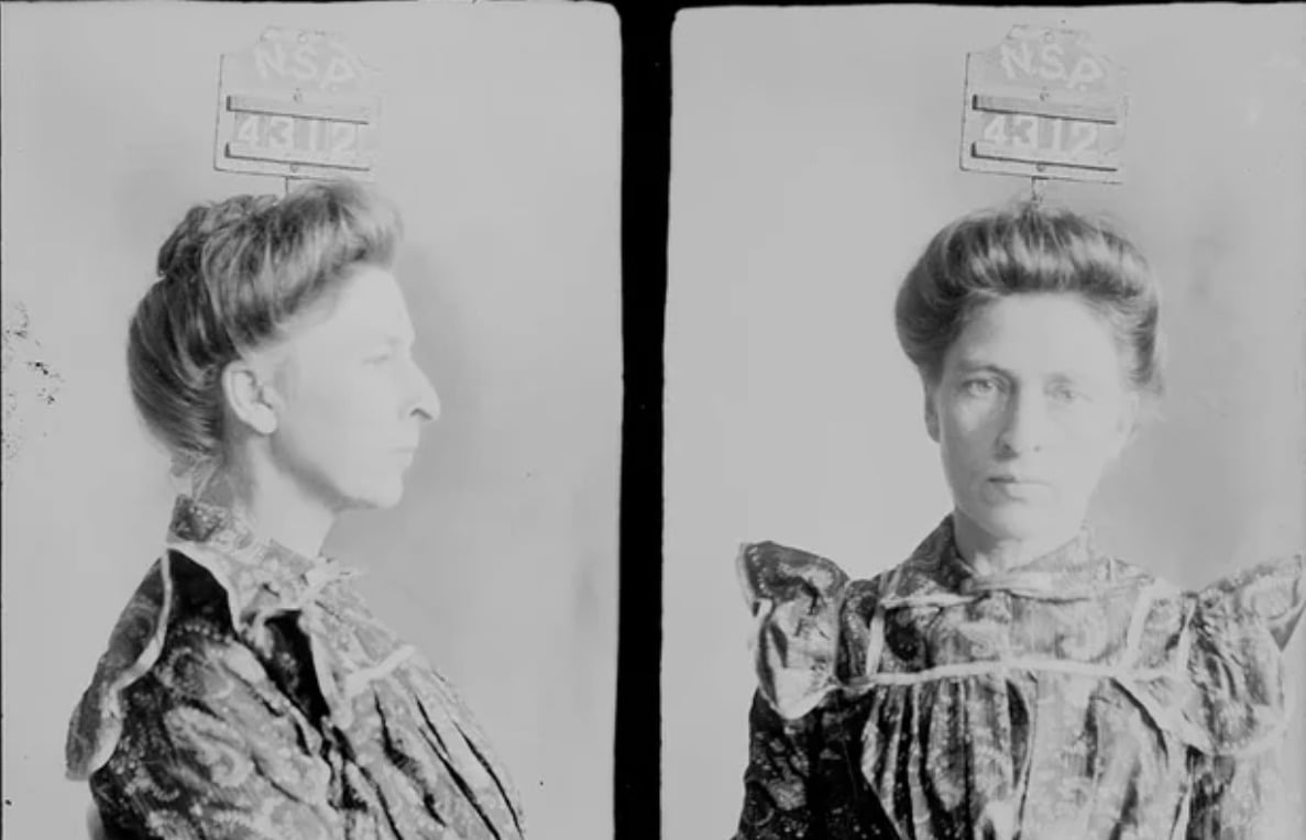 Black and white mugshot photo of a woman with light-colored hair styled up, wearing a patterned dress. The photo shows her in both profile and front-facing views with a sign labeled “NSW 4312” above her head.