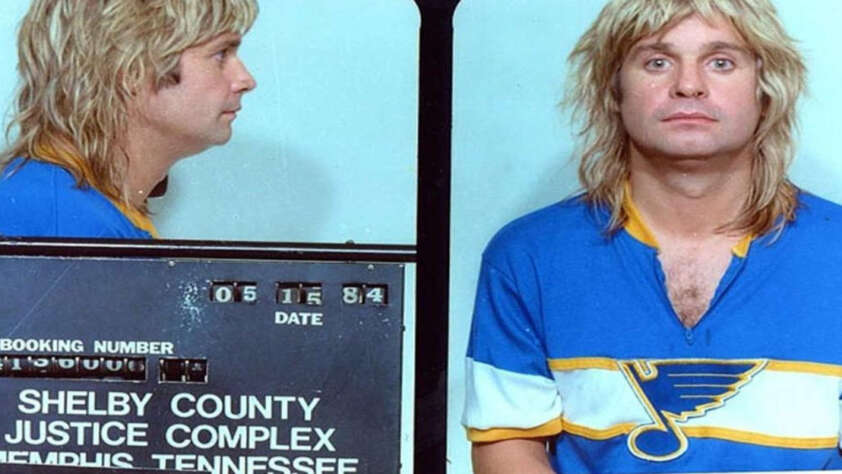 A man with long blonde hair wearing a blue St. Louis Blues shirt is shown in a mugshot with front and side views. The sign reads "Shelby County Justice Complex, Memphis Tennessee, 05-18-84.