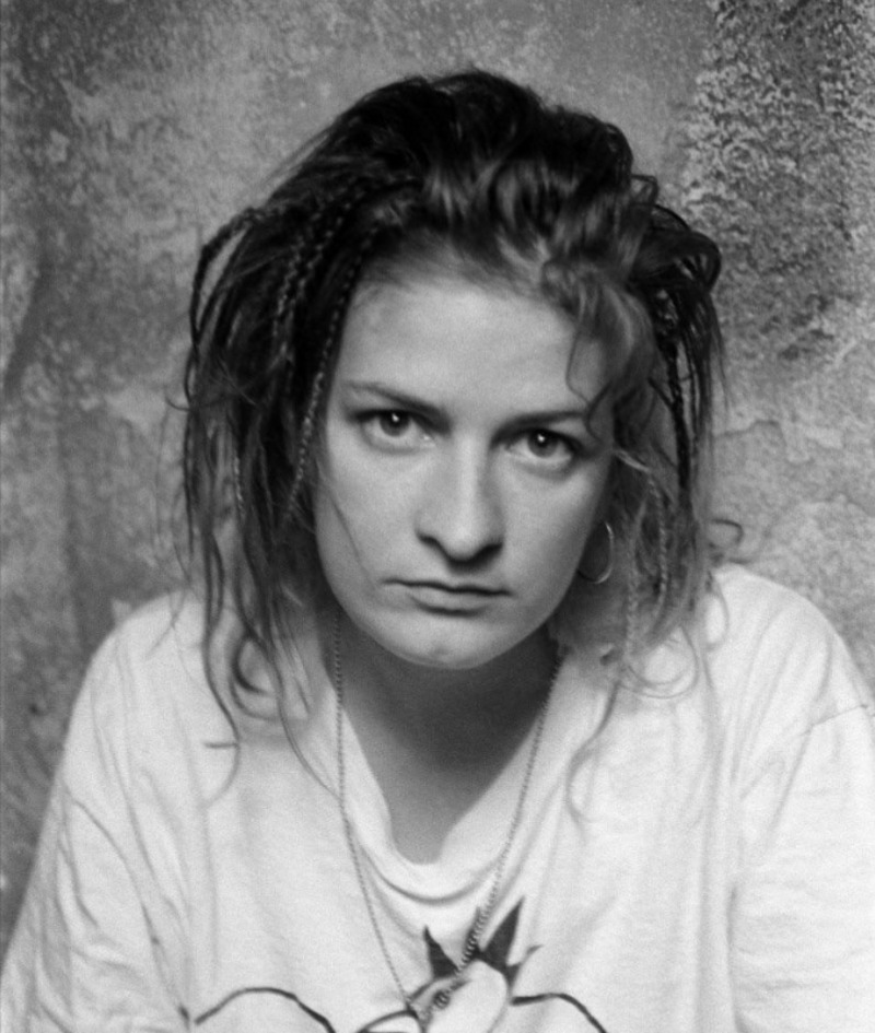 A young woman with loose, messy hair and small braids stares seriously at the camera. She is wearing a light-colored T-shirt and hoop earrings, with a textured wall in the background.