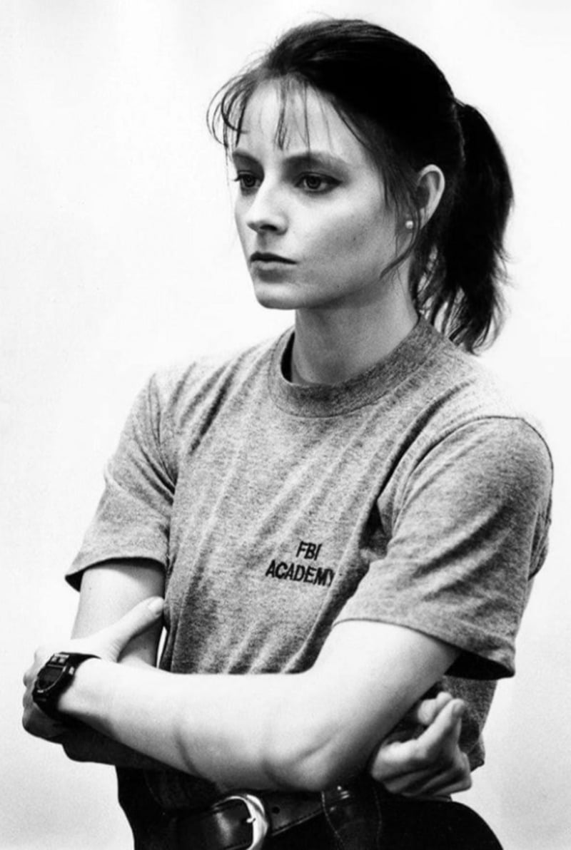 A woman with a serious expression stands with arms crossed, wearing a grey "FBI Academy" t-shirt and a watch, her hair tied back in a ponytail, against a plain light background.
