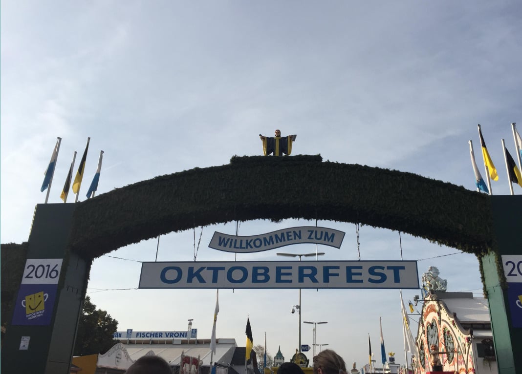 A large green archway decorated with flags and a "Willkommen zum Oktoberfest" sign welcomes visitors to Oktoberfest 2016 in Munich, Germany. A person stands atop the arch under a blue sky.