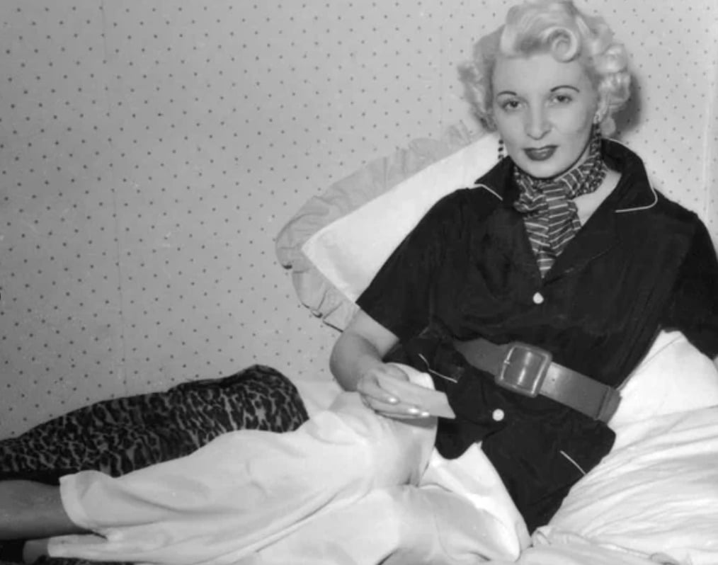 A woman with short, curled blonde hair reclines on a bed, wearing a dark blouse, wide belt, light-colored pants, and a striped scarf, with one leg crossed over the other. The wall behind her has a subtle dotted pattern.