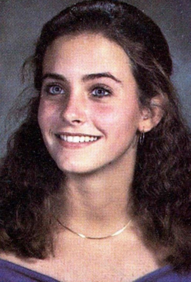 A young woman with wavy brown hair, blue eyes, and light skin smiles. She wears a purple top, a gold necklace, and small hoop earrings, posed in front of a gray backdrop, as in a school portrait.