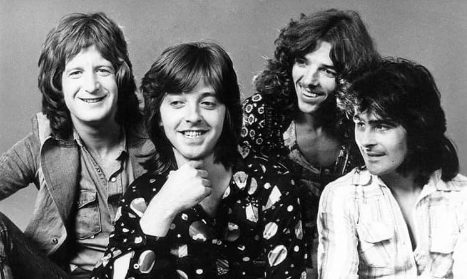 Four young men with long hair sit close together, smiling and looking relaxed. The photo is black and white, and they wear casual, patterned 1970s-style shirts against a plain background.