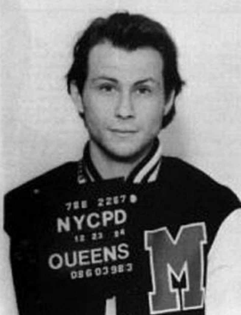 Black and white mugshot of a young man with short hair wearing a varsity jacket with a large "M" on it, holding a sign that reads "NYCPD QUEENS.