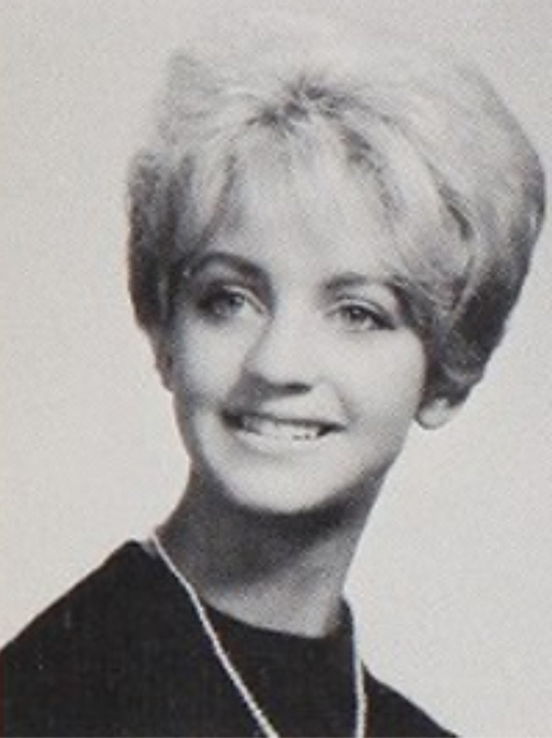 Black and white portrait of a young woman with light-colored, styled hair, wearing a dark top and a necklace, smiling and looking slightly to the side.